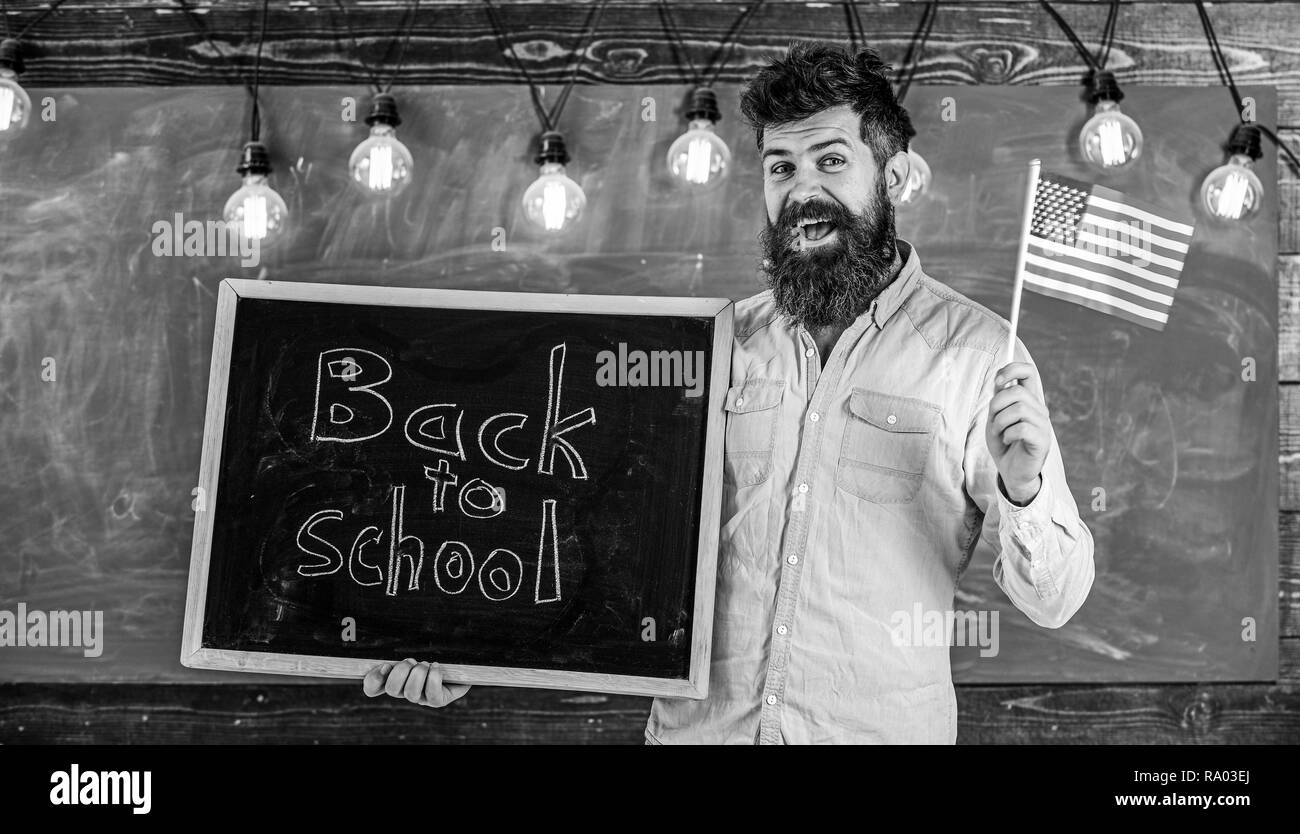 American school concept. Teacher holds blackboard with written phrase back to school and flag of USA. Man with beard on smiling face welcomes to american school, chalkboard on background. Stock Photo
