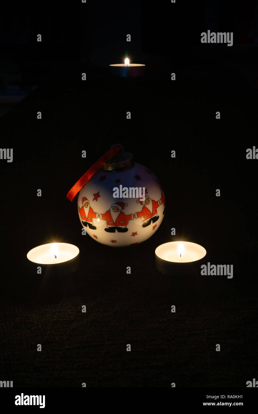 A decorative Christmas bauble depicting three Santa Clauses with two lit candles next to it against black background - Christmas concept Stock Photo