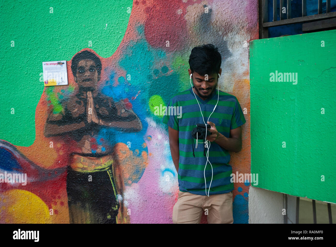 15.04.2018, Singapur, Republik Singapur, Asien - Ein junger Mann steht vor einem bunten Wandbild im Stadtteil Little India und schaut auf sein Smartph Stock Photo