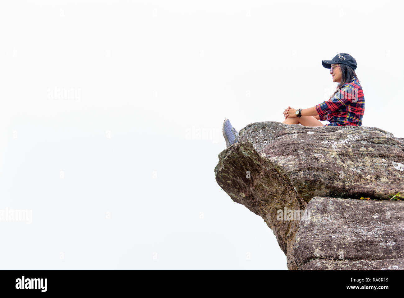 Asian woman sitting on a rock near a cliff on a white fog background looking forward with a happy smile. Stock Photo