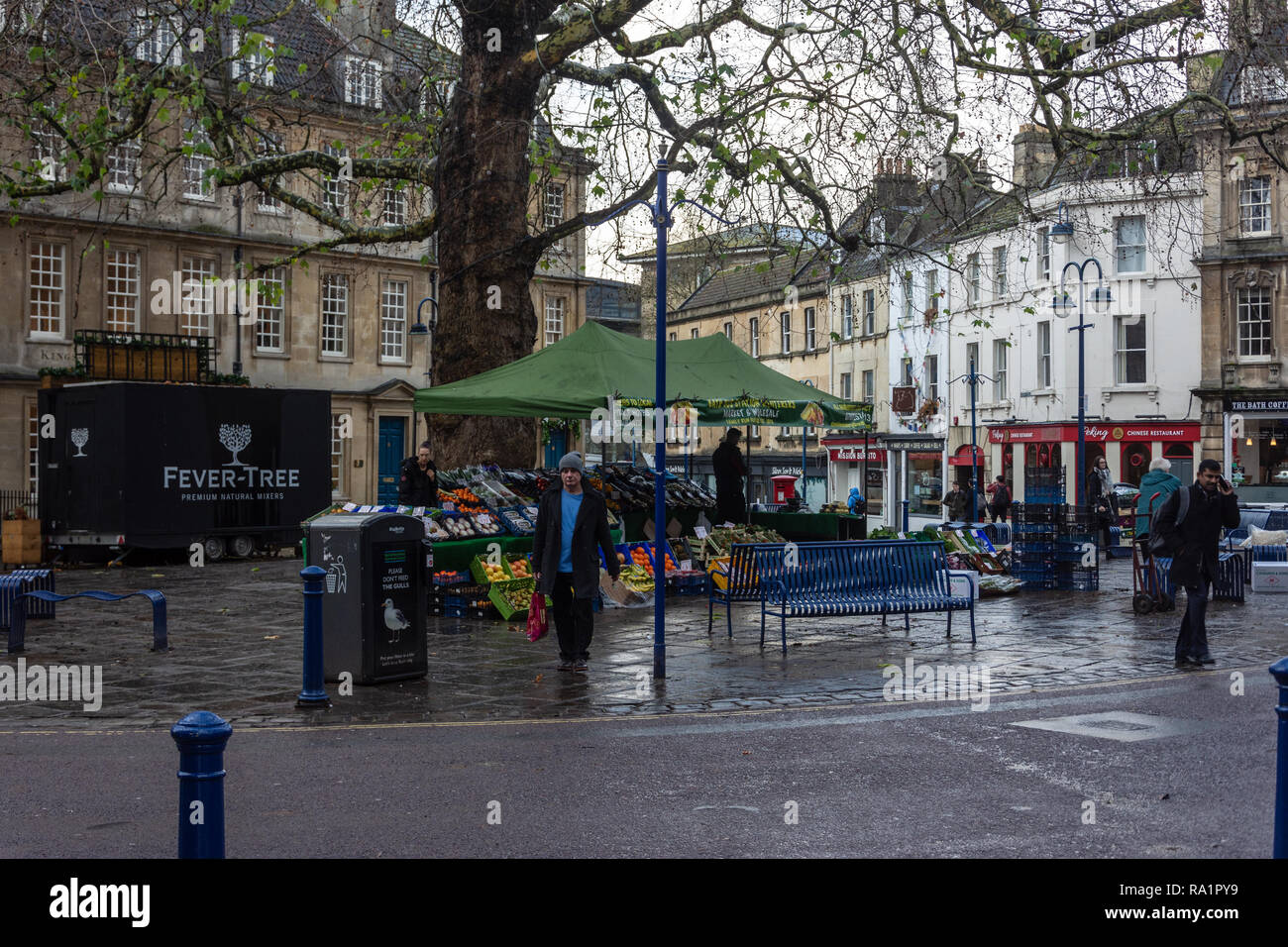 A person with a carrier bag walking away from a fruit and veg market stall set in Kingsmead Square in the city of Bath UK on a wet winter morning Stock Photo