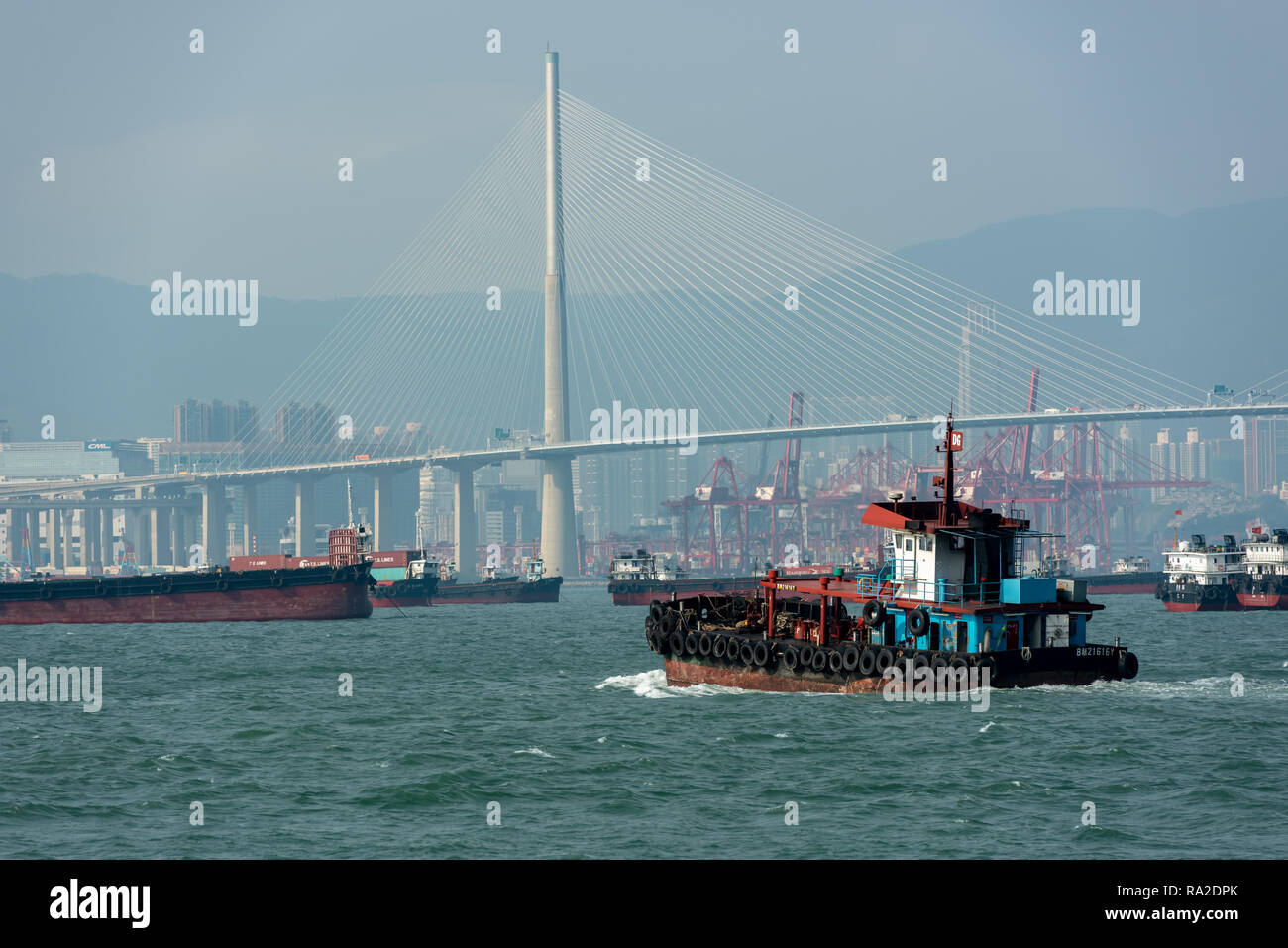 A colourful tugboat navigates the busy shipping lanes around Stonecutters Bridge and Tsing Yi's container terminal in Hong Kong. Stock Photo