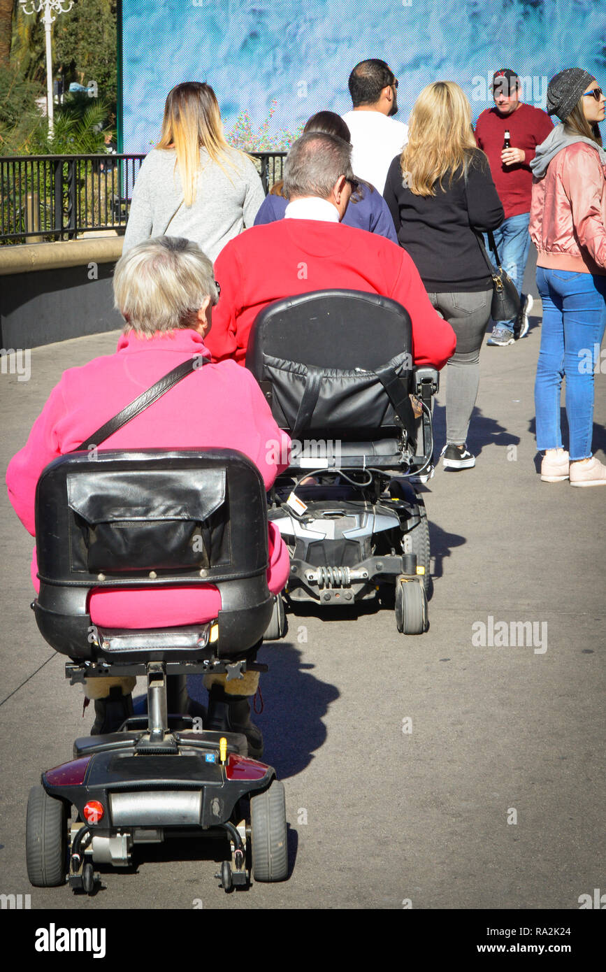Rear view of A man and a woman, both in wheel chair style scooters joining the crowds on the sidewalk while sightseeing in the USA Stock Photo
