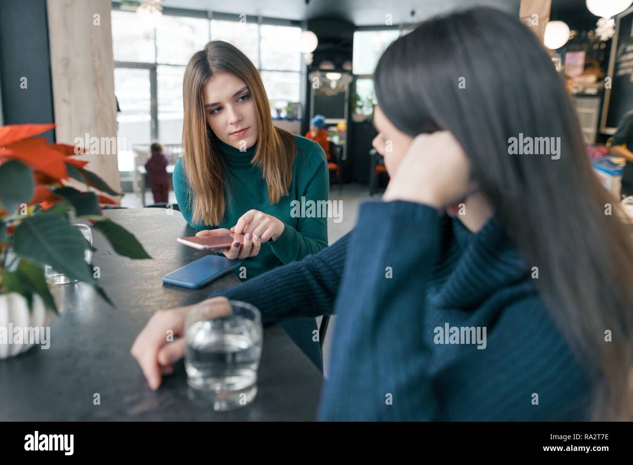 Talking young women, girls sitting in winter cafe smiling and talking, drinking clean water in glass and using mobile phone, red poinsettia christmas Stock Photo