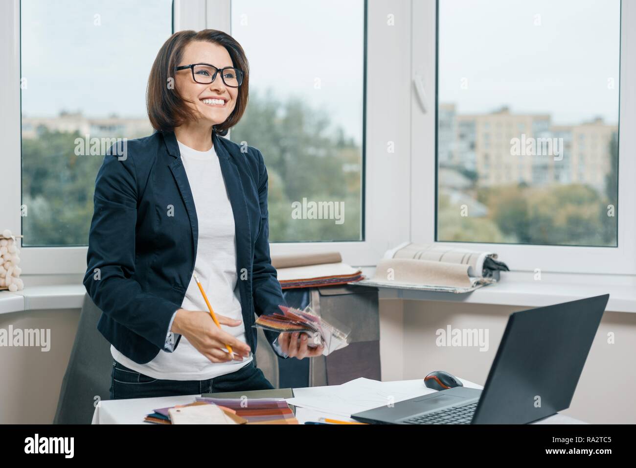 Female decorator interior designer at workplace with fabric samples, laptop. Smiling female in jacket, glasses looking to the side while standing near Stock Photo