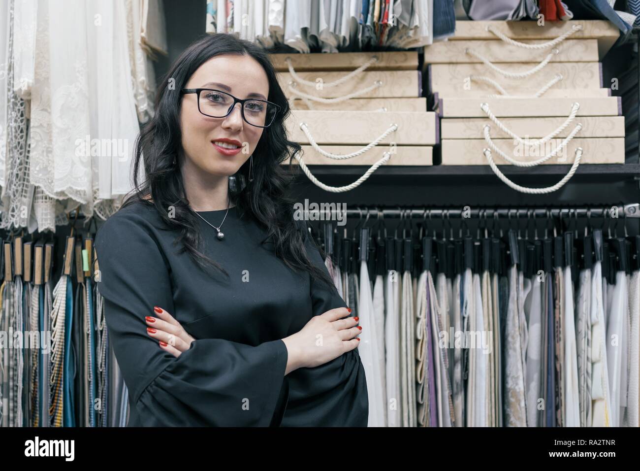Confident businesswomen owner of a small business fabric store, woman with glasses smiling with folded hands at the workplace, background fabric sampl Stock Photo