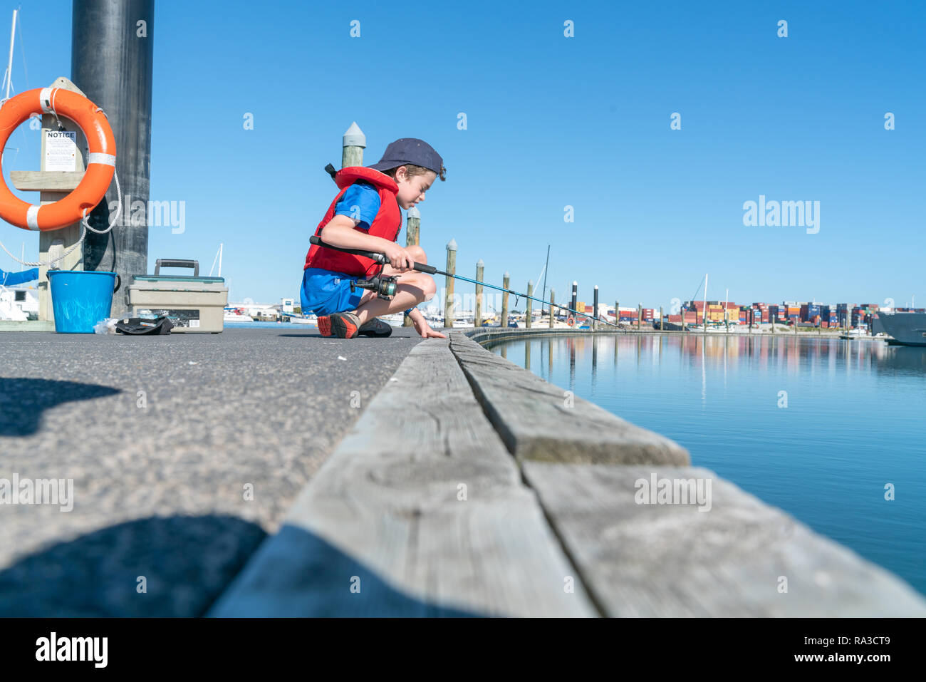 Boy sits on dock fishing wearing red life-jacket looking over edge into Tauranga harbour in marina New Zealand. Stock Photo