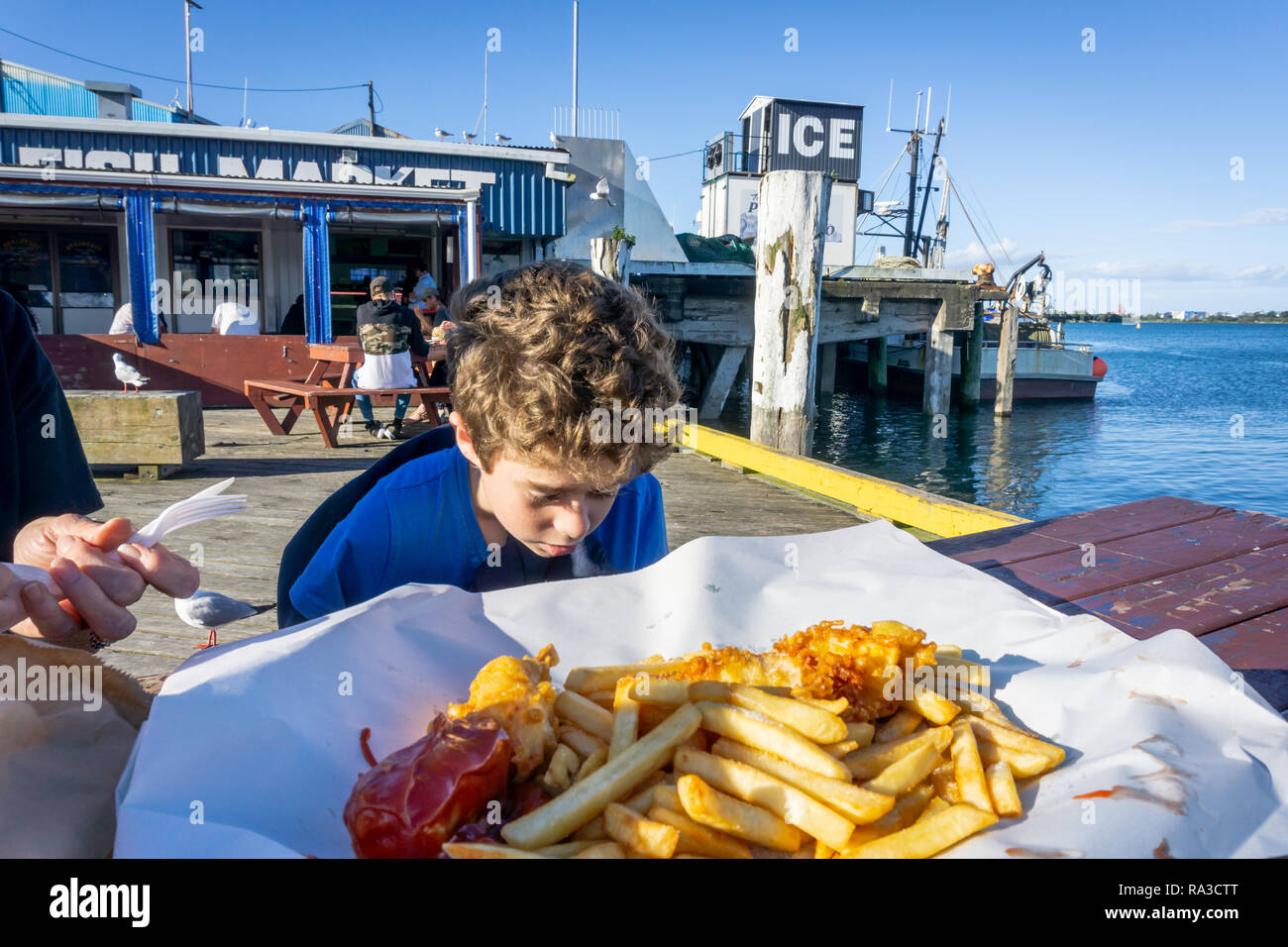 TAURANGA, NEW ZEALAND DECEMBER 20 2018; Small boy in blue jersey outside fish market with srpead of fish and chips on paper looks before eating. Stock Photo