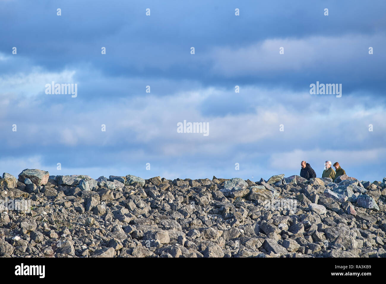Three people walk along the path behind the rock pile of the dam wall at Rutland Water reservoir in the Midlands of England on a cloudy winter day. Stock Photo