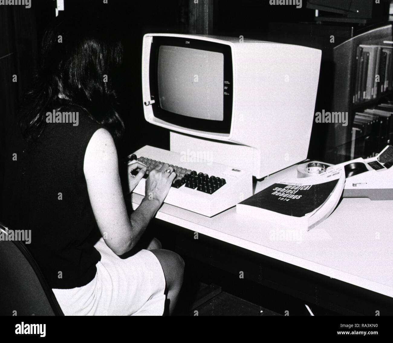 An operator is sitting in front of an IBM; 1974 Medical Subject Headings is next to the computer Stock Photo