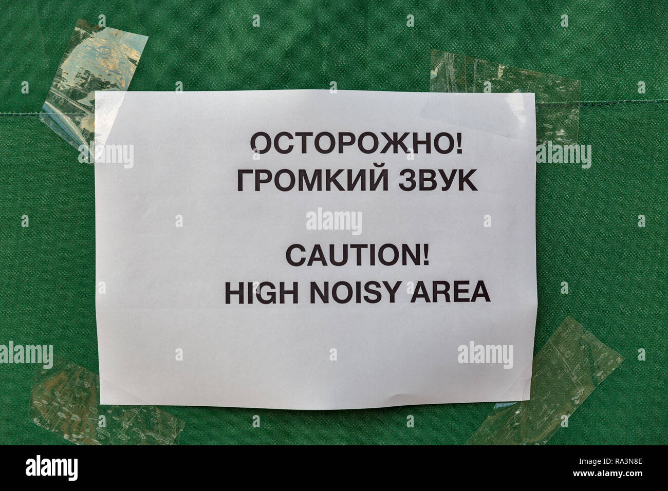 inscription in Russian and English on a white piece of paper taped by scotch on green tent background: Caution! High noisy area Stock Photo