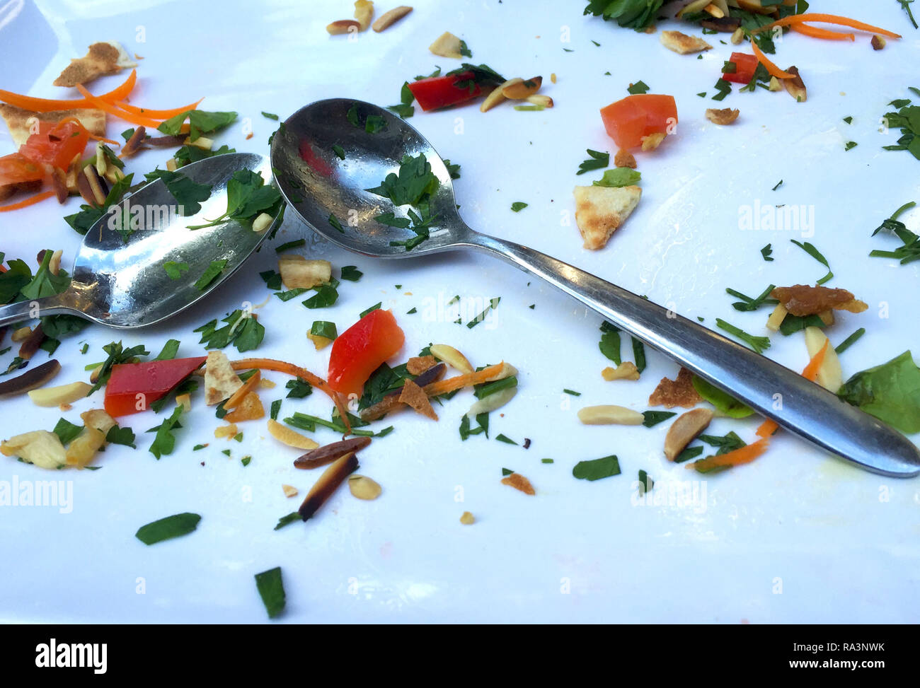 The remians of a meal are on a white plate. This includes tomato, pinenuts, carrot, slivered almonds, parsley and bread. Two silver spoons are on teh  Stock Photo