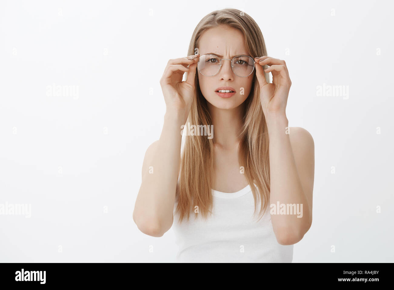 Girl cannot read sign putting on glasses squinting and frowning as looking focused at camera, trying understand what written having problems with sight standing clueless and intense over gray wall Stock Photo