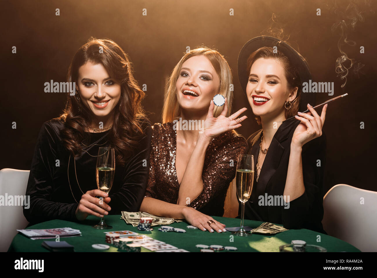 laughing attractive girls with glass of champagne, cigarette and poker chips sitting at table in casino Stock Photo