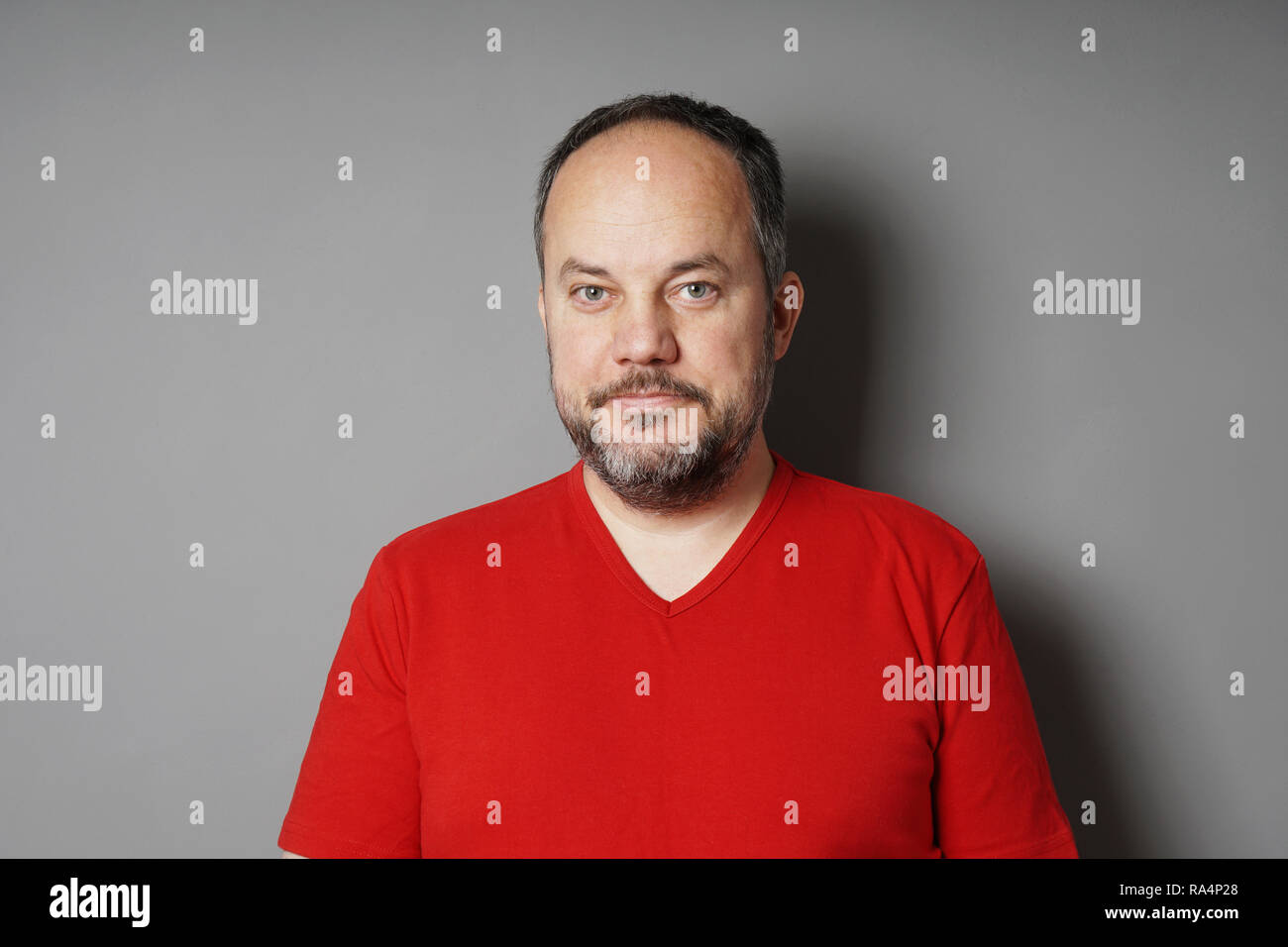 middle aged man in his 40s wearing red t-shirt with short dark hair and graying beard smirking - gray wall background with copy space Stock Photo
