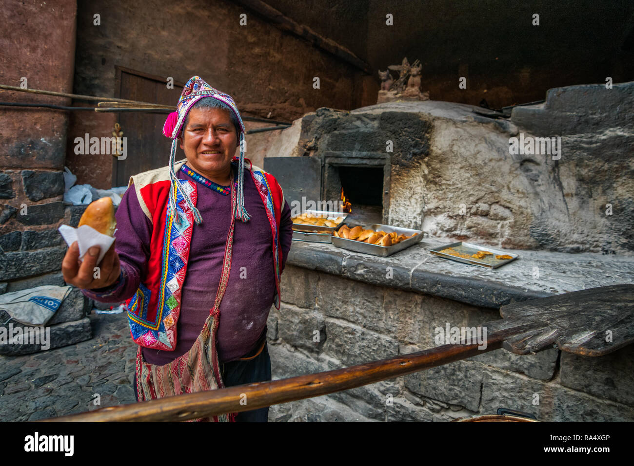 PISAC MARKET, PERU - SEPTEMBER 2018 : Man baker in traditional Peruvian cloth and hat, holding fresh baked empanada, standing near outdoor stone stove Stock Photo