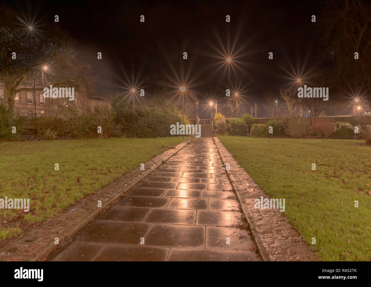 A wet dawn at  Tower Gardens in York.  A path runs straight ahead and the lawns are covered in autumn leaves.  Street lamps light up the steps. Stock Photo