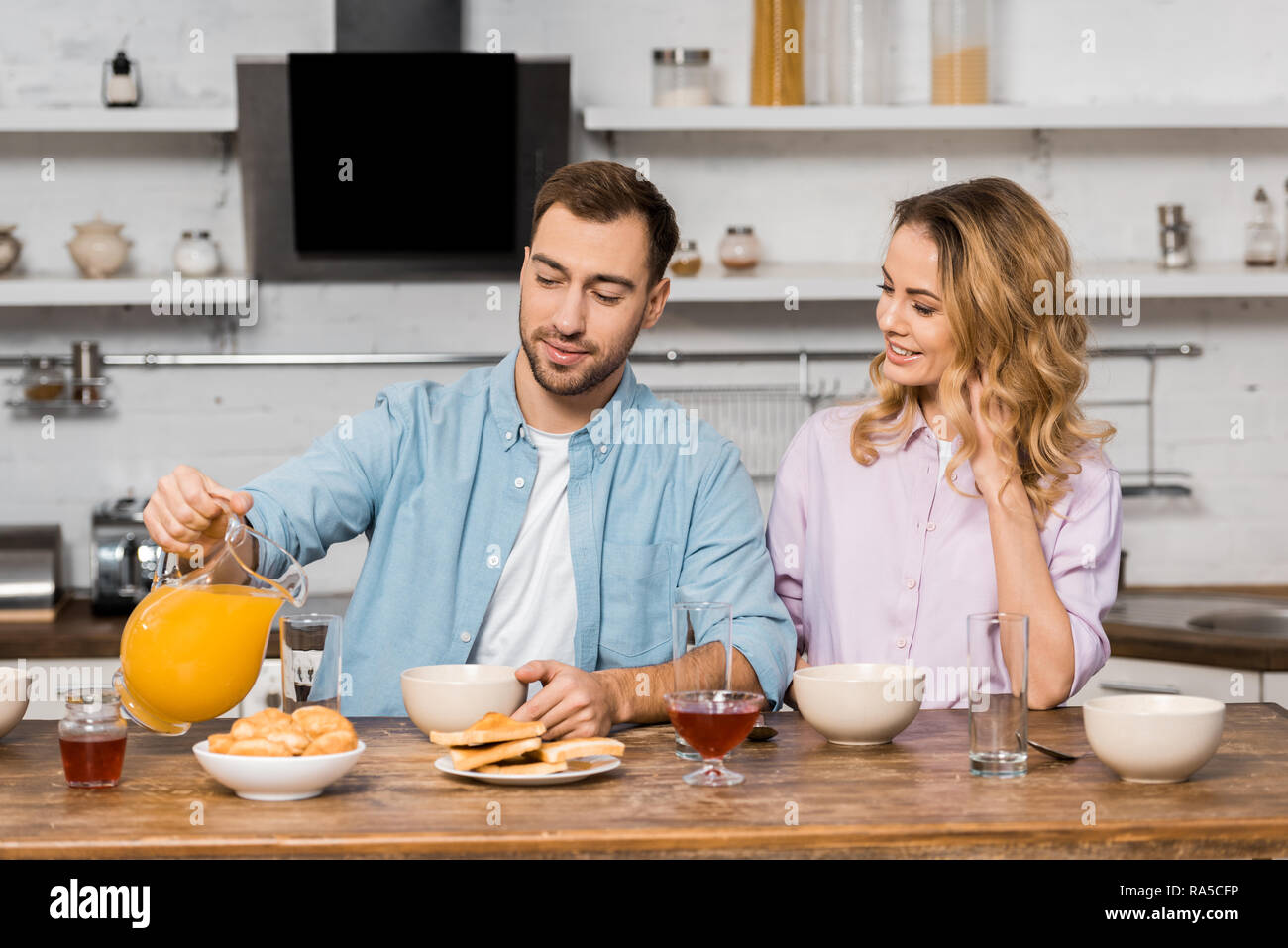 smiling woman looking at handsome husband pouring orange juice in glass on kitchen table Stock Photo