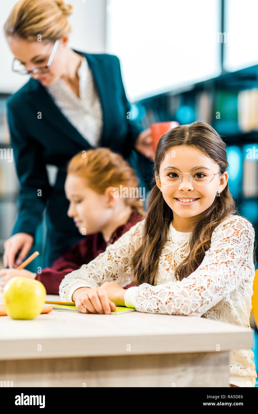 cute schoolkid in eyeglasses smiling at camera while studying with classmate and teacher in library Stock Photo