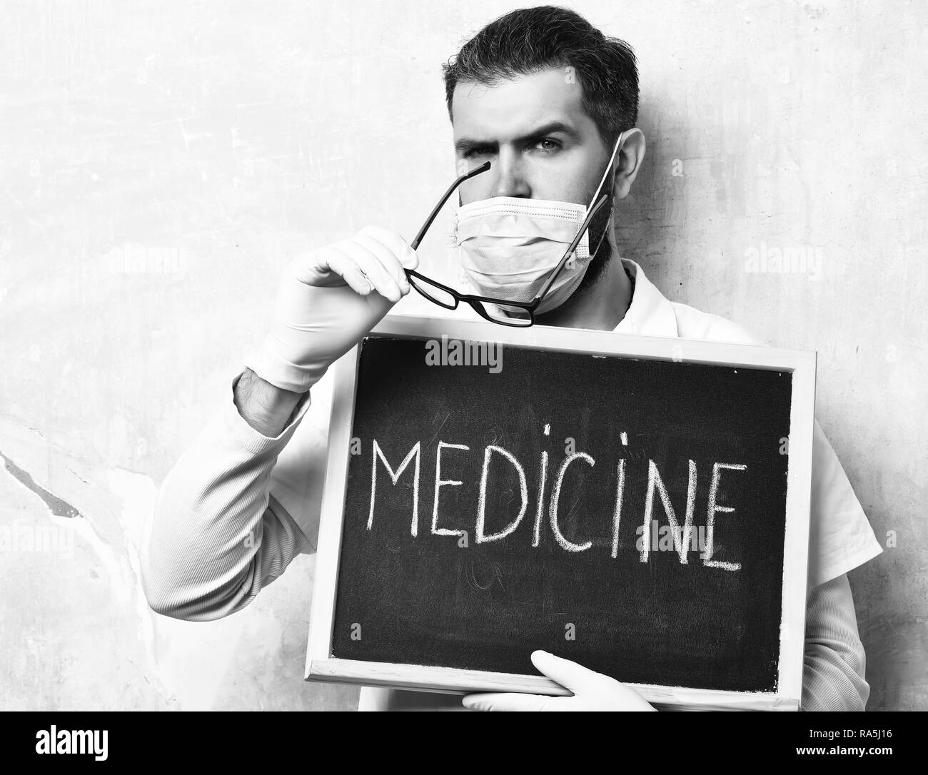 Bearded man, long beard. Brutal caucasian doctor or unshaven hipster, postgraduate student in medical gown, mask holding board with Medicine inscription on texture studio background. Medicine concept Stock Photo