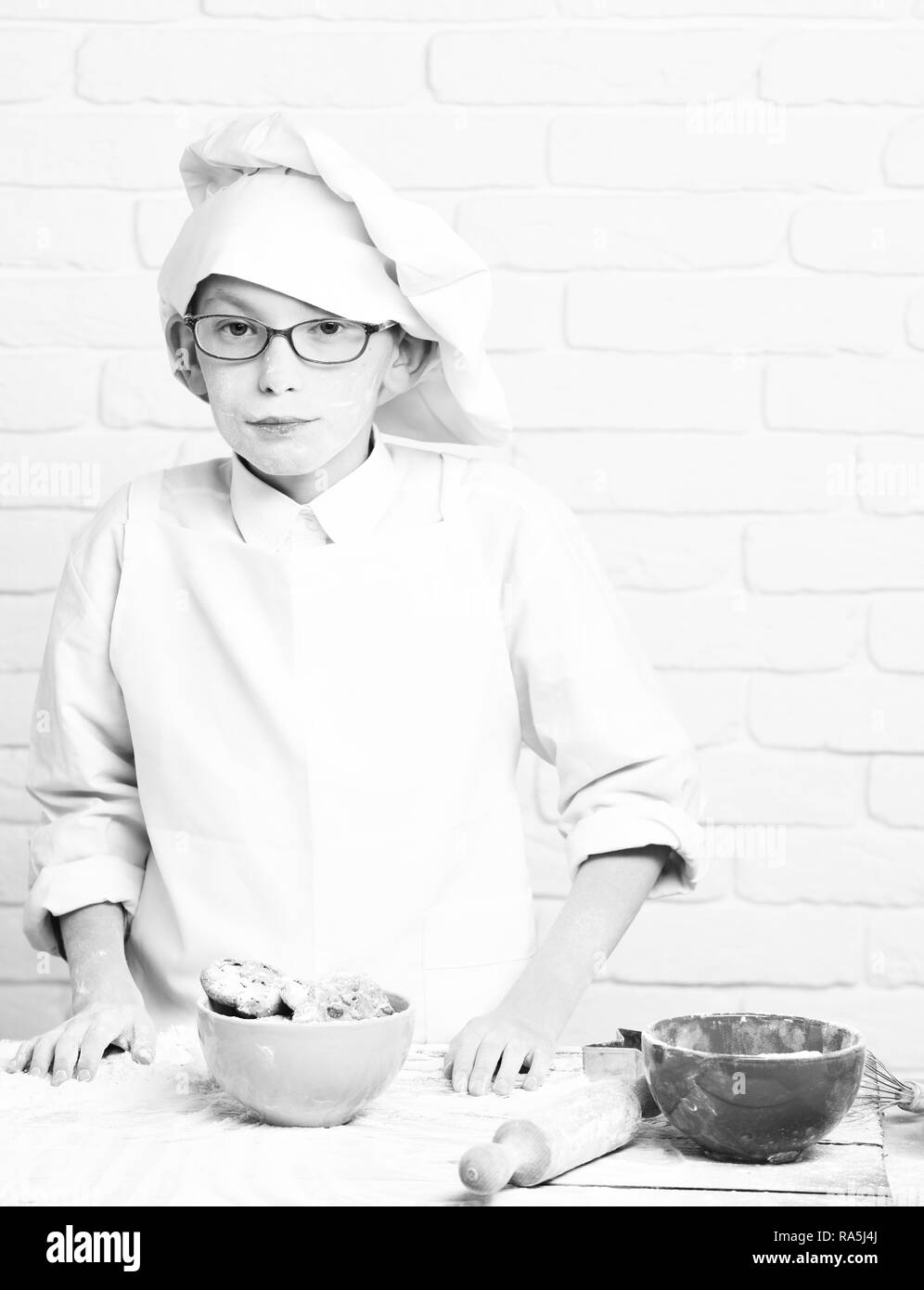 young boy cute cook chef in white uniform and hat on stained face flour with glasses standing near table with rolling pin and colorful bowls with chocolate cookies on brick wall background. Stock Photo