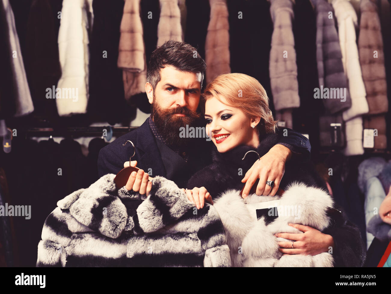 Woman with smiling face in black fur coat with bearded man. Luxury shopping concept. Couple in love holds white sable and chinchilla fur coats. Man with strict face and woman with coats in fur shop. Stock Photo