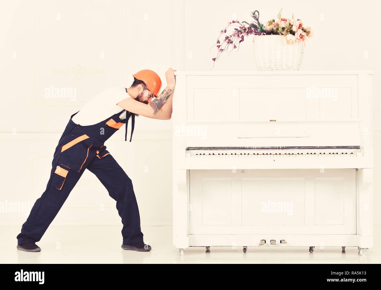 Man with beard worker in helmet and overalls pushes, efforts to move piano, white background. Heavy loads concept. Courier delivers furniture, move out, relocation. Loader moves piano instrument. Stock Photo