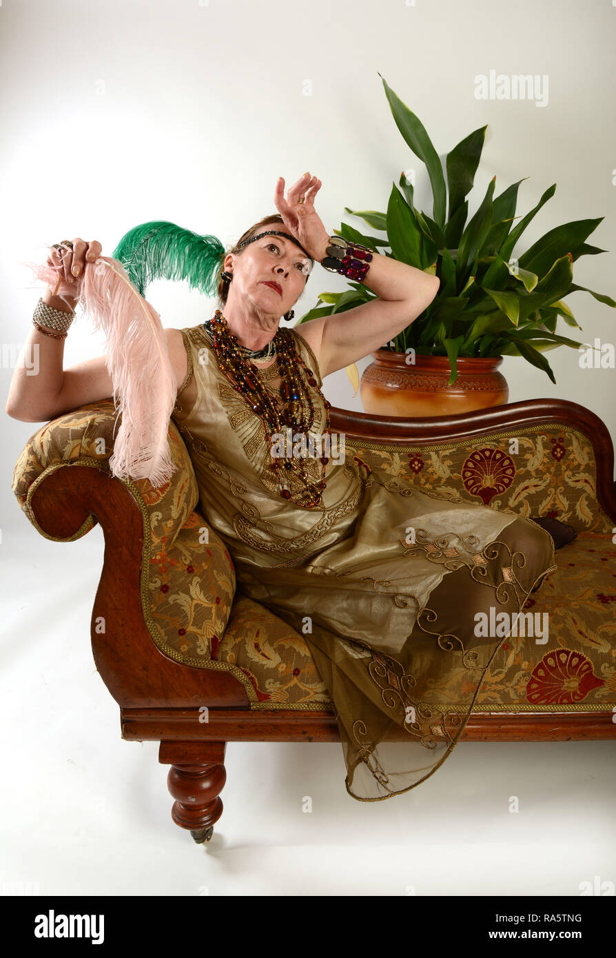 Senior middle aged woman posing as 1920's flapper dressed fainting lady on chaise longue Stock Photo