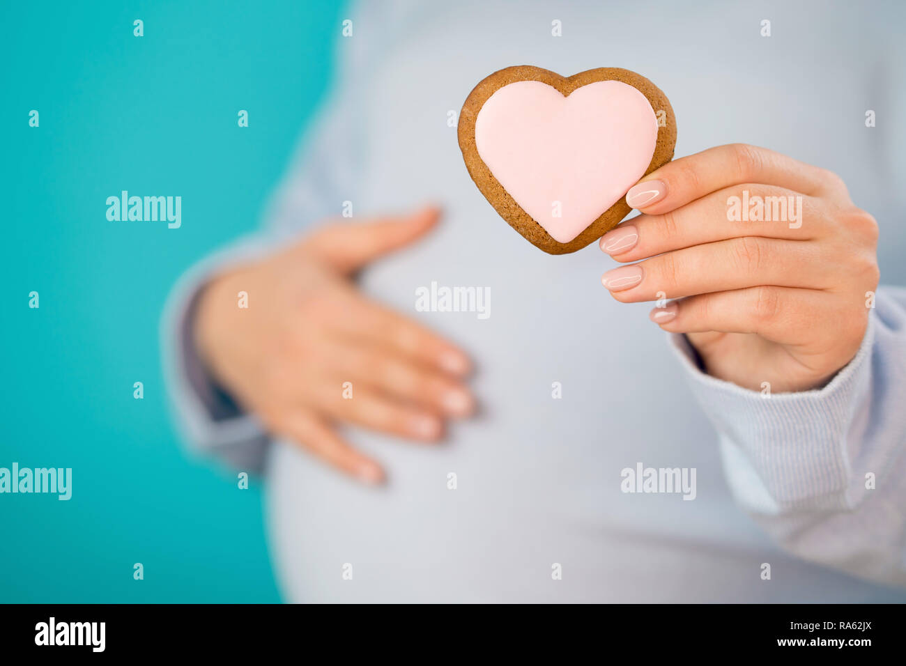 Pregnant woman holding heart-shaped cookie on tummy belly background. Young girl in blue wearing expecting baby. Maternity, motherhood, pregnancy, love concept. Stock Photo
