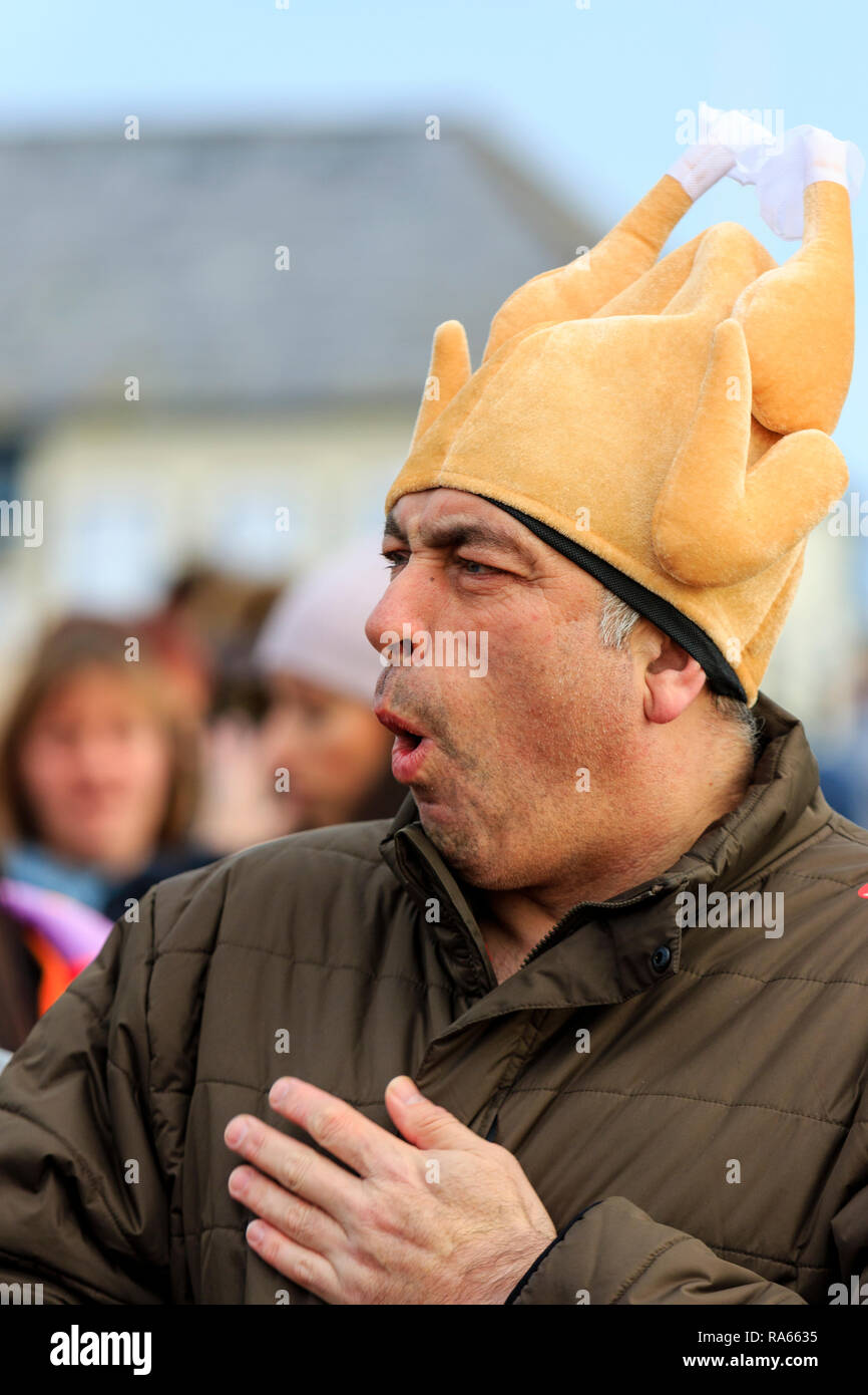 Close up, side view, head and shoulders of mature man, male, talking and gesturing with his hands. Wears a yellow chicken hat. People in background. Stock Photo