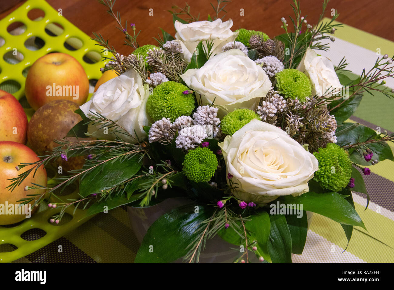 Beautiful bouquet of flowers with white ranunculus buttercup, green carnation and small purple and white flowers on wooden table. Light green hole dotted plate with fruits apple and pear Stock Photo