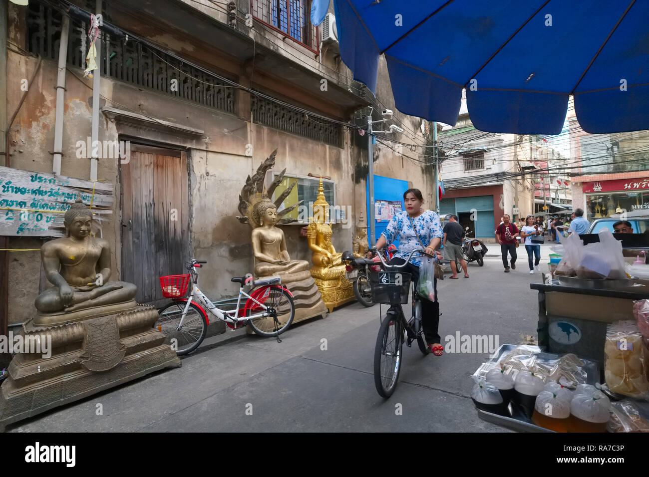 A woman cycles past Buddha statues placed near a factory producing Buddha statues and other religious objects; Bamrung Muang Rd., Bangkok, Thailand Stock Photo