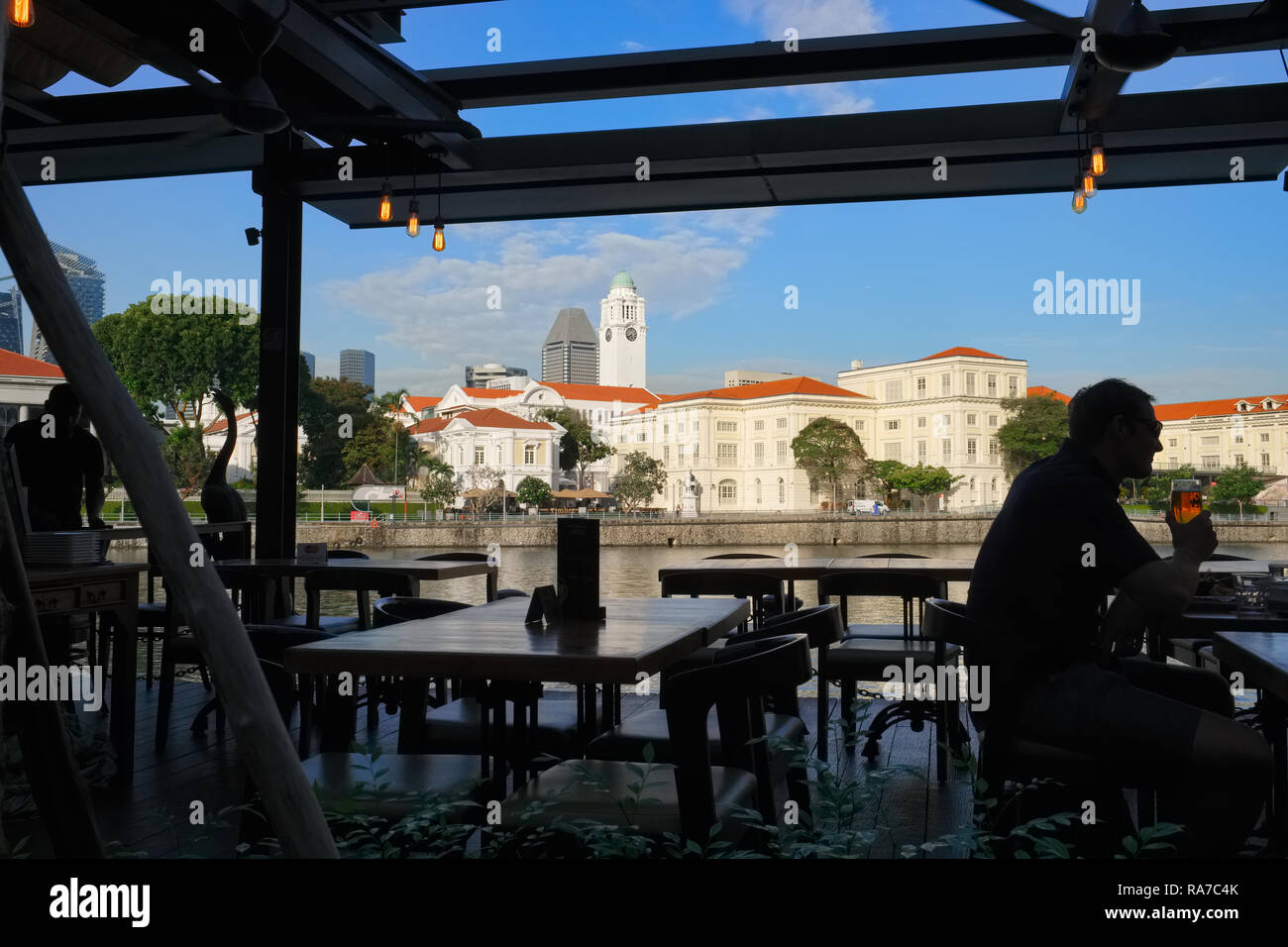 A man drinks a beer in a bar at Boat Quay by the Singapore River, Singapore; b/g: Empress Place and the clocktower of Victoria Theatre & Concert Hall Stock Photo