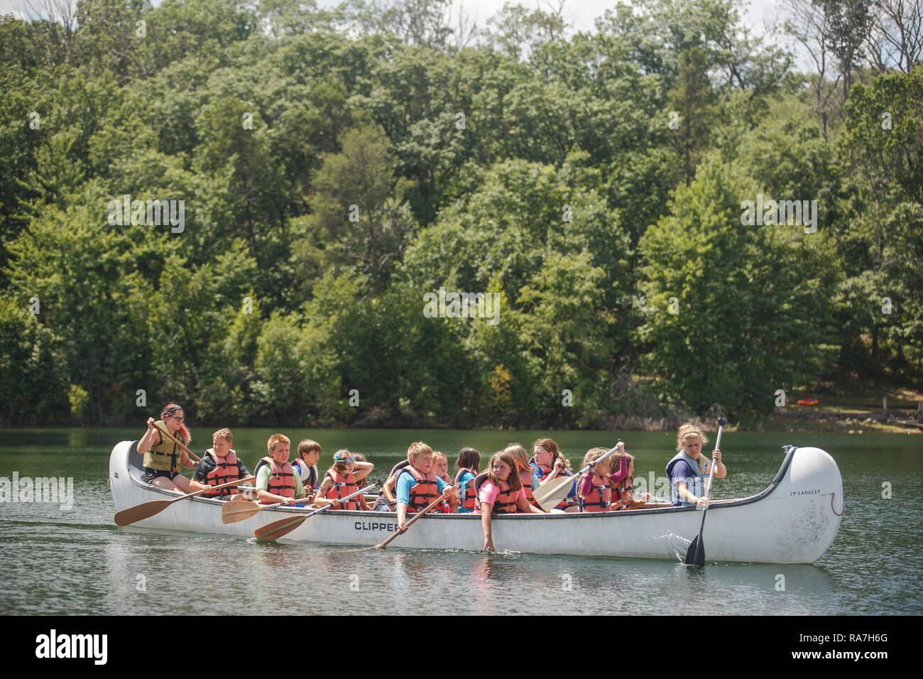 Many young children wearing life preservers fill a large canoe that paddles in a lake during their summer camp. Stock Photo
