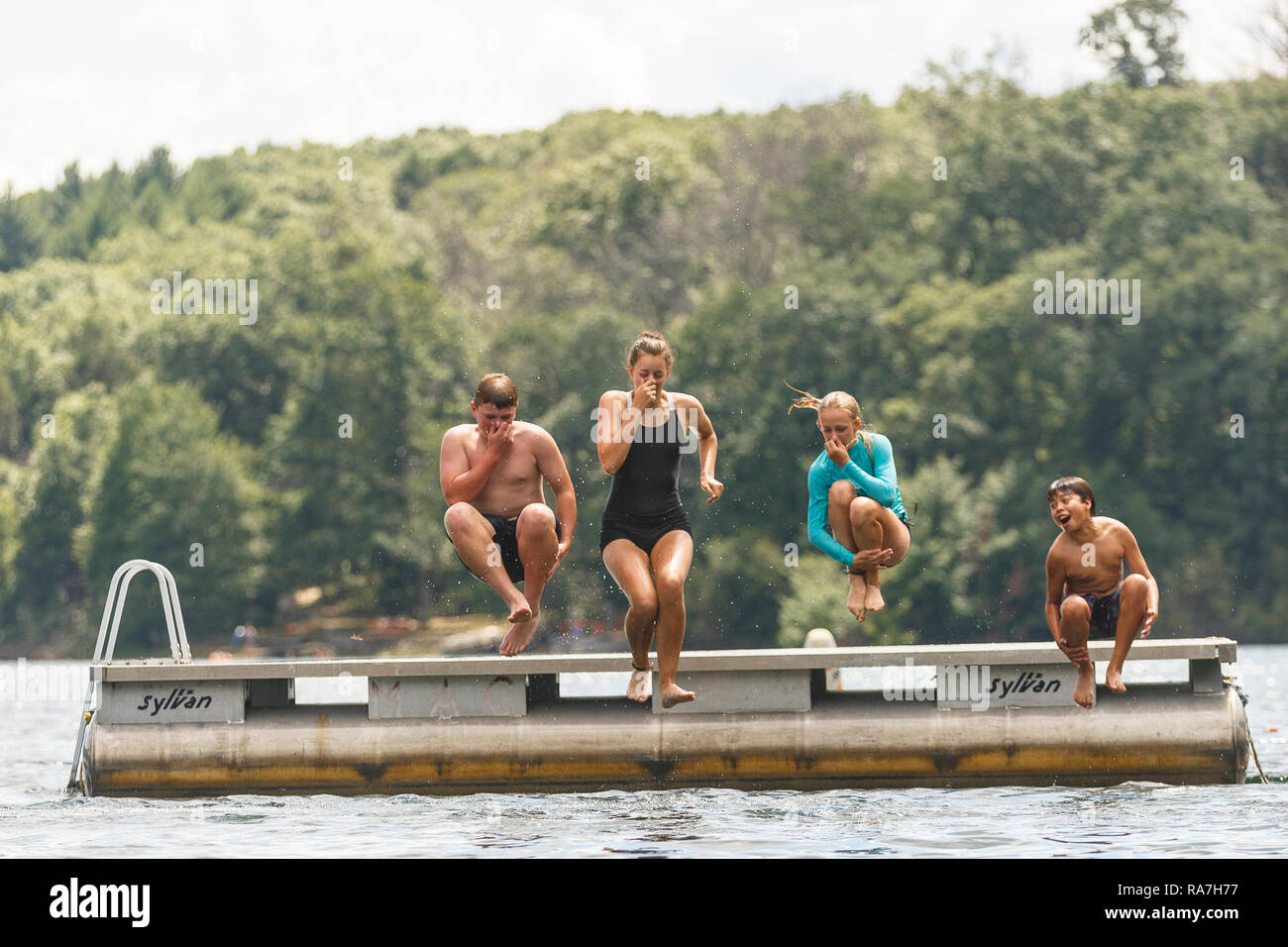 A group of young kids jump off of a raft that is floating in the middle of a lake at a summer camp Stock Photo