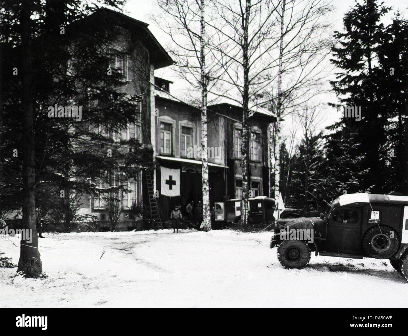 Front exterior view of a country villa that has been converted into a medical clearing station. Snow covers the front yard. Two U.S. Army trucks sit in front. The rear door of the truck closest to the villa is opened, and military personnel hover between the truck and the building. Stock Photo