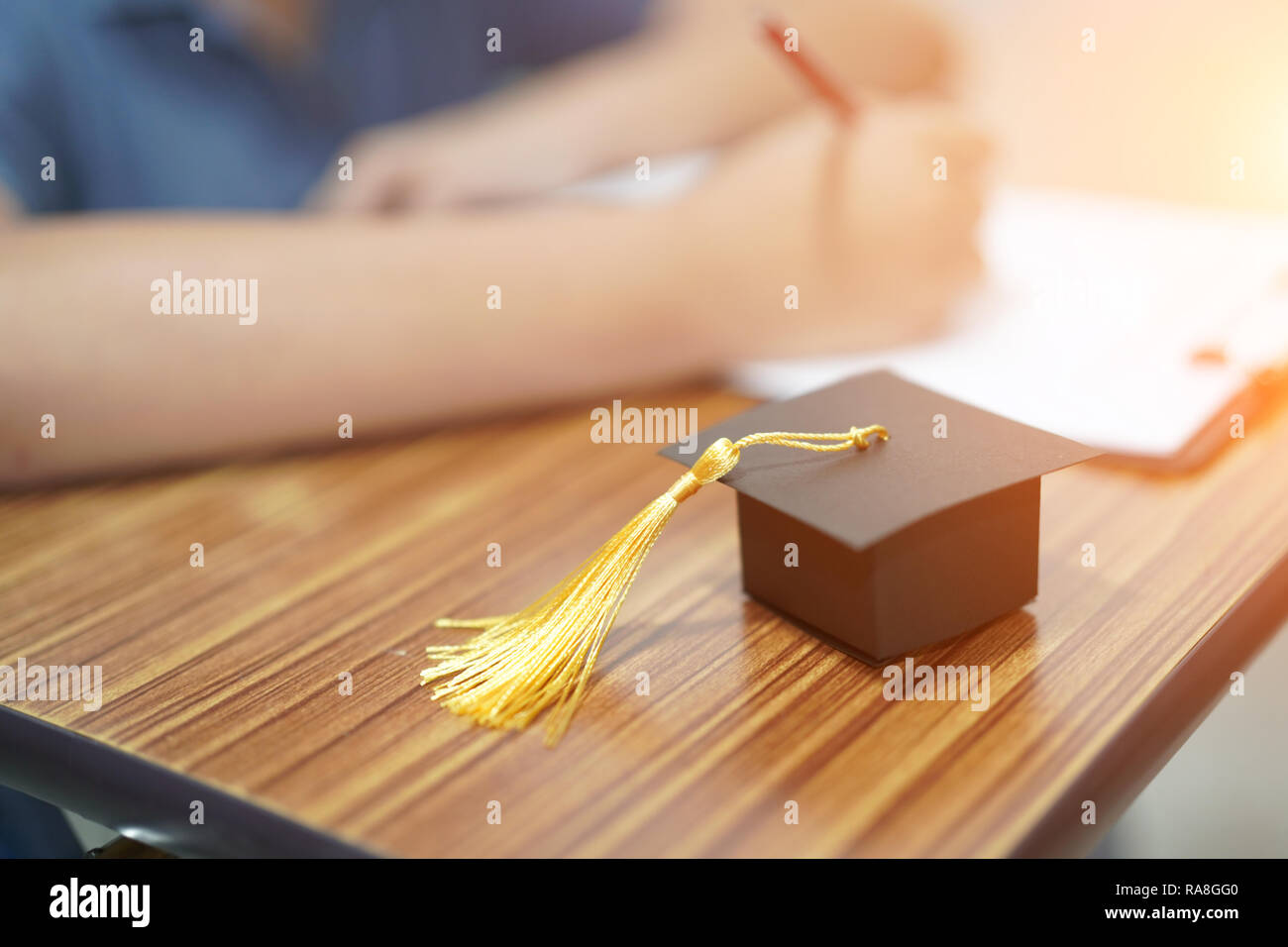 Asian children young kid write answer in a quiz examination test study learn with graduation gap hat on wooden table in education school. Stock Photo