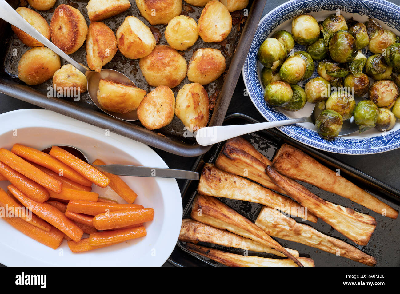 Roast potatoes, brussel sprouts, carrots and parsnips in baking trays and bowls with spoons on a slate background Stock Photo