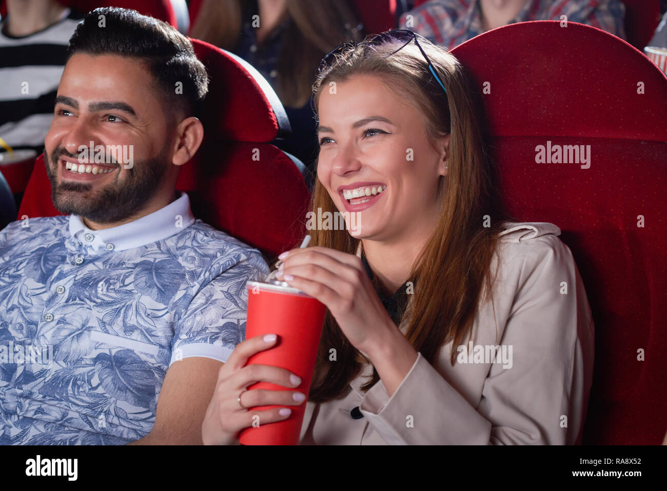 Positivity couple of blonde in gray and Arabian man in blue shirt drinking and smiling spending time in cinema. Students having fun, when looking at screen in modern cinema hall. Stock Photo
