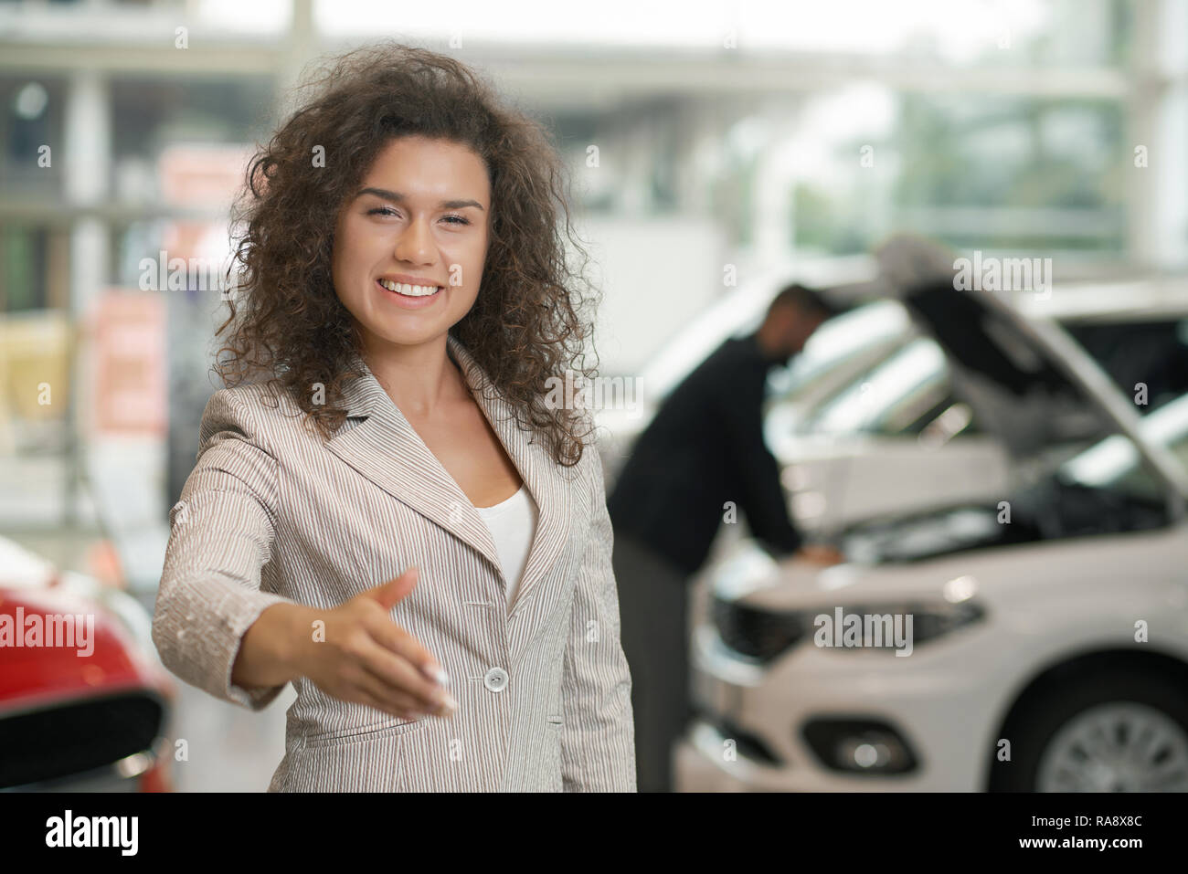 Female car dealer smiling and holding hand for shaking. Happy manager making deal in car showroom. Beautiful with curly hair looking at camera, posing. Client of car center observing vehicle behind. Stock Photo
