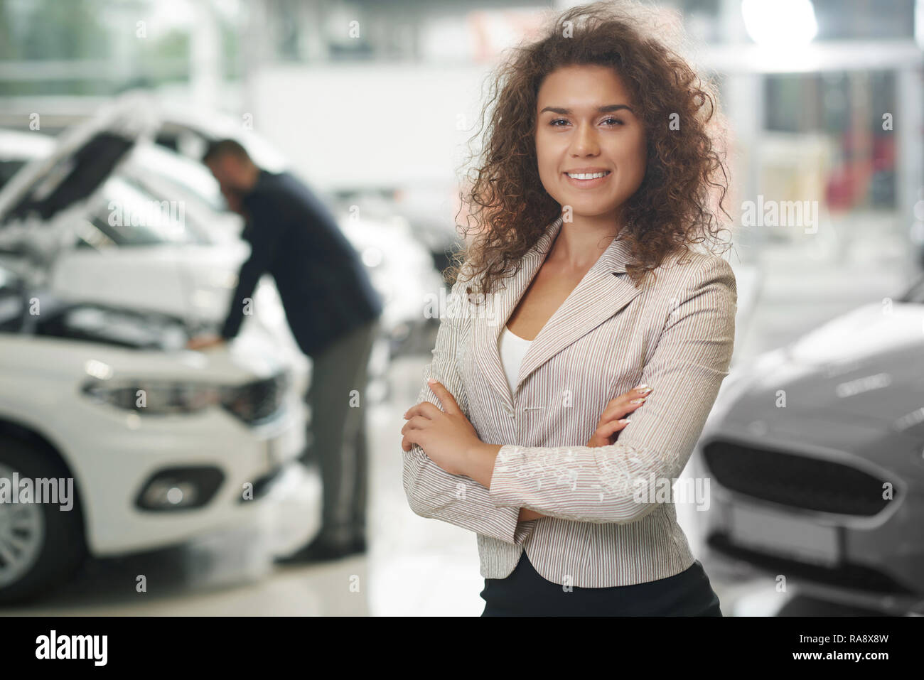 Gorgeous female manager posing in car dealership. Charming woman with curly hair looking at camera, smiling. Car dealer working in car showroom. Man observing car behind. Stock Photo