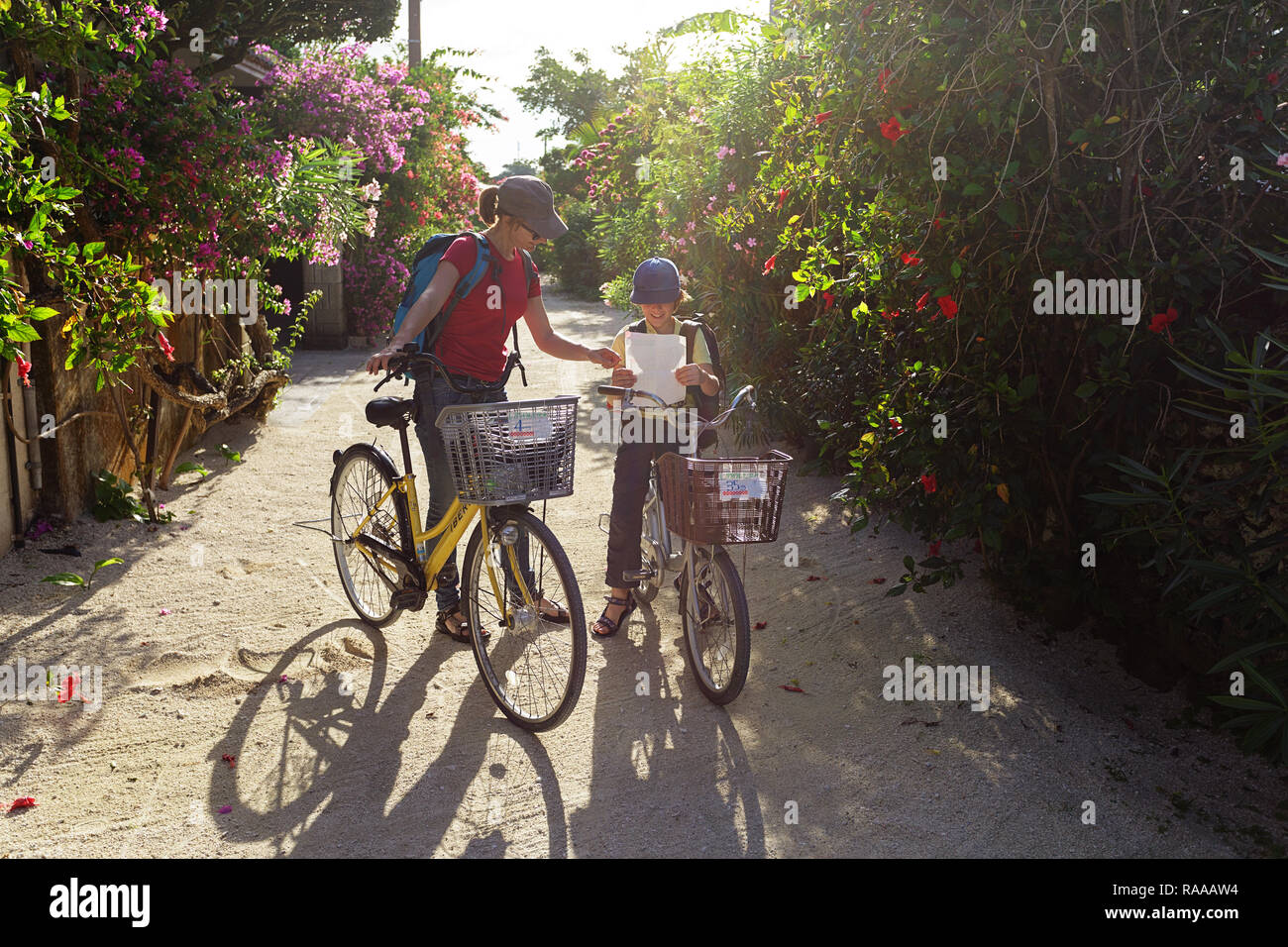 Mother and son on bicycles reading a map exploring traditional village of Taketomi on streets covered with hibiscus island in Okinawa,Japan Stock Photo