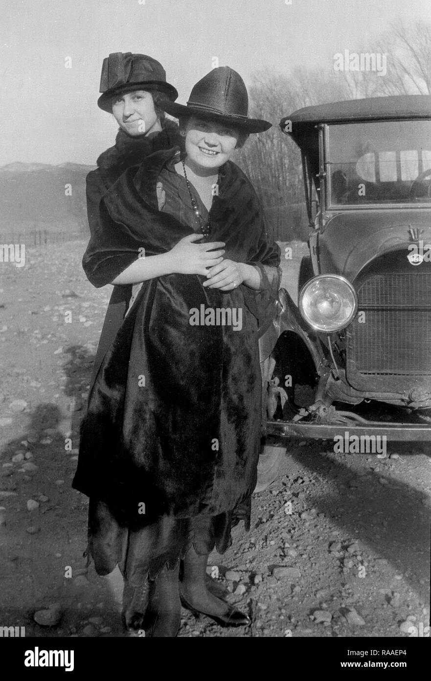 Two women hug while posing with their automobile along a Colorado road, ca. 1920. Stock Photo