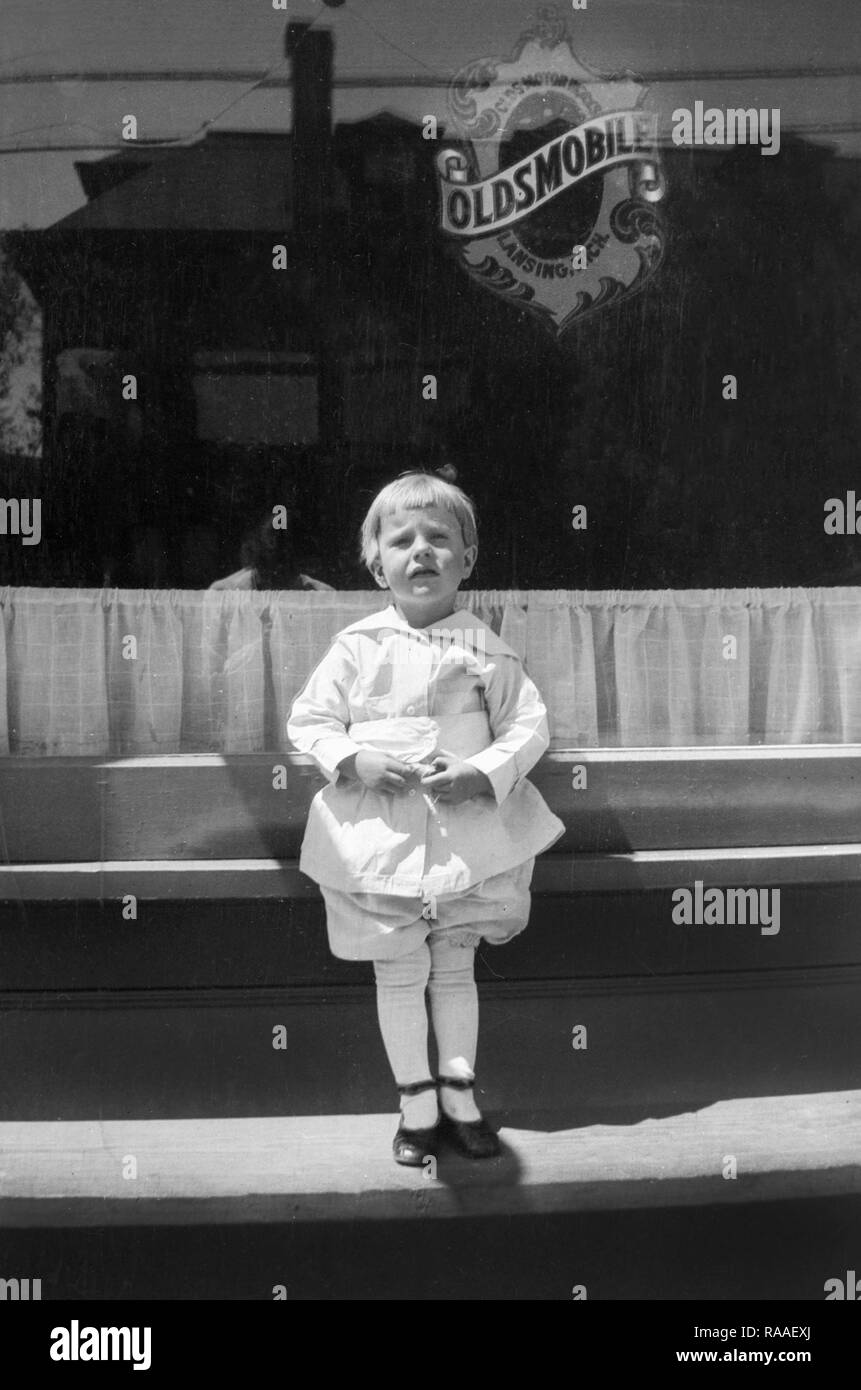 A young child stand outside a Oldsmobile dealer, ca. 1925. Stock Photo