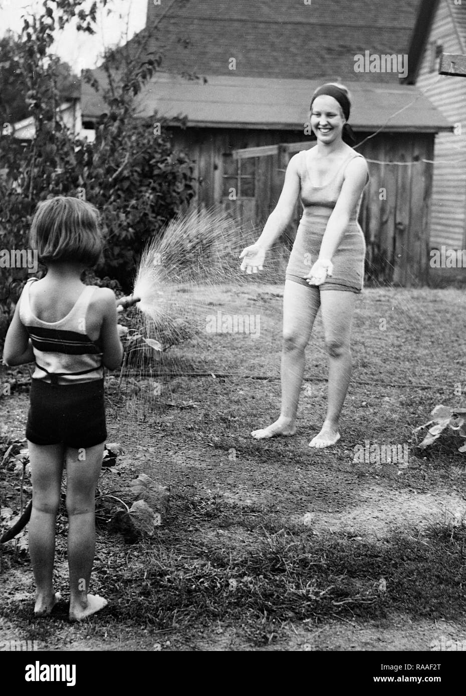 A young woman is cooled off by her young sister with the garden hose, ca. 1925. Stock Photo