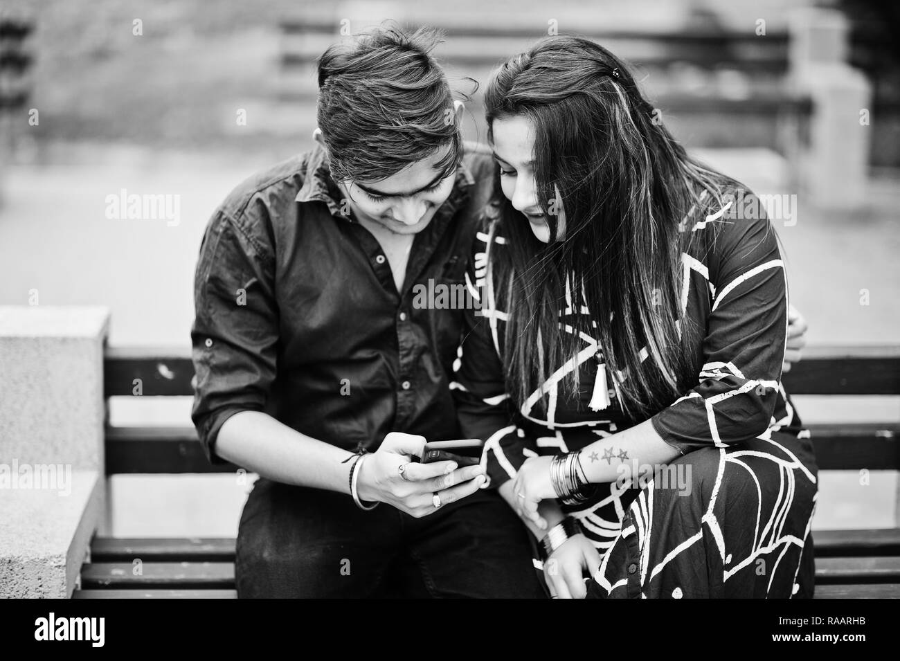 Indian couple posed outdoor, sitting on bench together and looking at mobile phone. Stock Photo