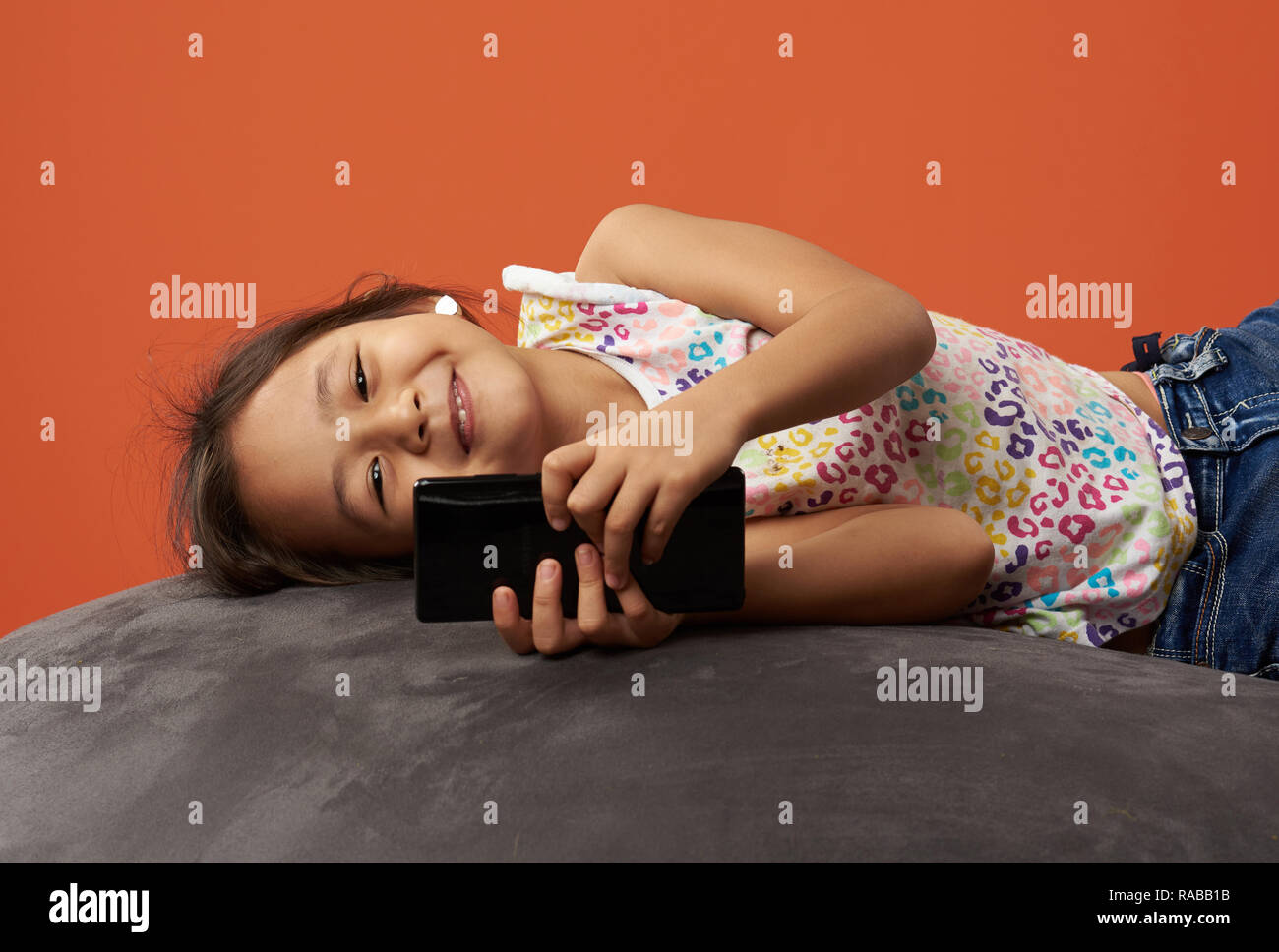 Asian kid smiling with phone in her hands. Kid on a bean bag holding phone. Stock Photo