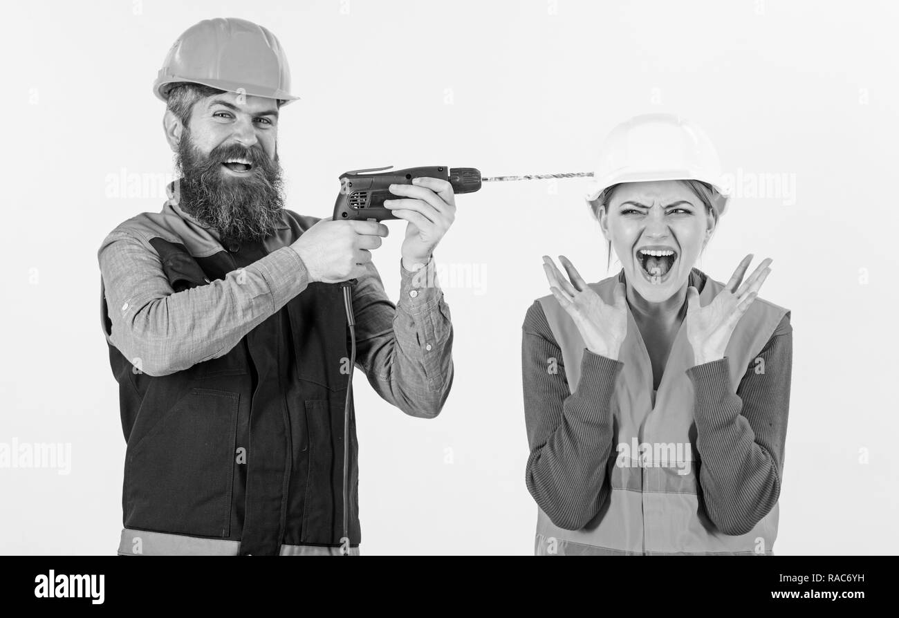 Husband annoying his wife. Man with happy face drills head of woman, white background. Builder, repairman makes hole in female head. Woman on scared face in helmet, hard hat. Annoying repair concept. Stock Photo