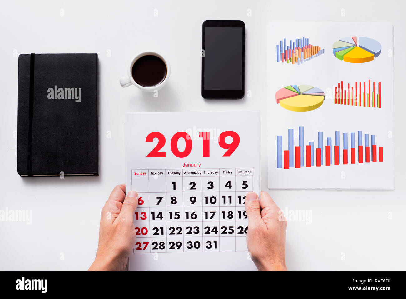 high angle view of a caucasian man sitting at his office desk observing a calendar of 2019, next to some charts, a cup of coffee, a smartphone and a n Stock Photo
