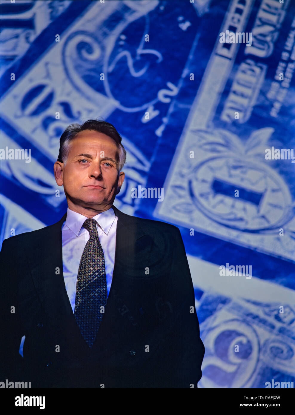Mature businessman or banker standing in front of a paper currency background with symbolism of greed, corruption and deceit on his face Stock Photo